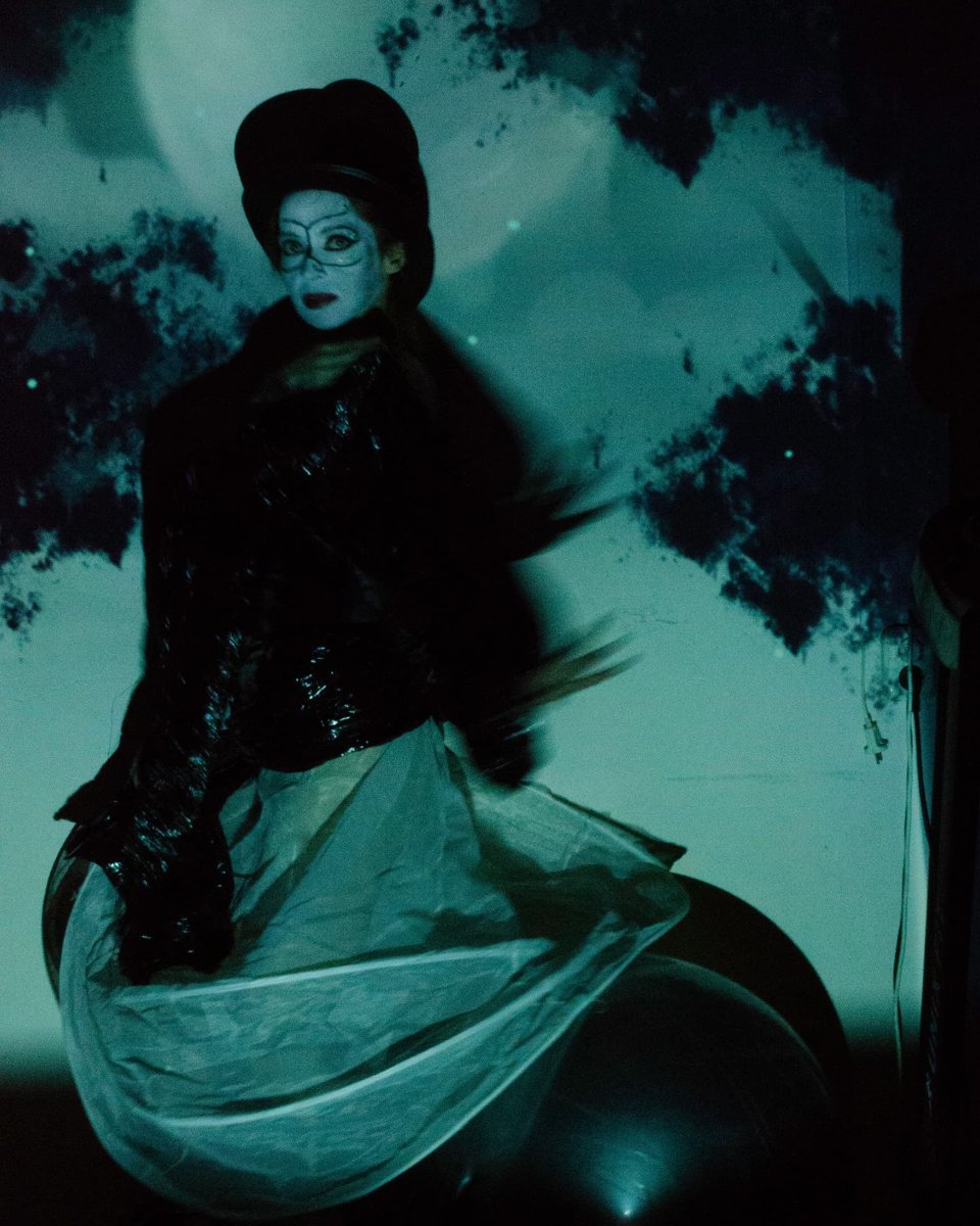 Video-art still: a masked figure bathed in teal projected foliage